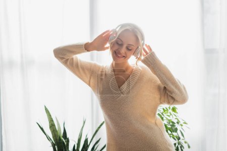 glückliche junge Frau mit drahtlosen Kopfhörern, die in einer modernen Wohnung Musik hört 