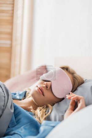 blonde woman in pink sleeping mask and blue pajama resting in bed 
