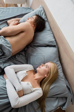 top view of shirtless man sleeping under blanket near embarrassed blonde woman after one night stand 