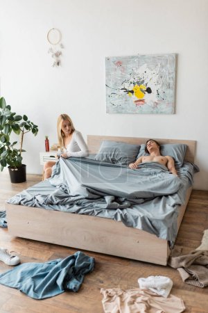 Blonde Frau sitzt auf Bett und zieht Decke, während hemdloser Mann nach einem Nachtstand schläft  