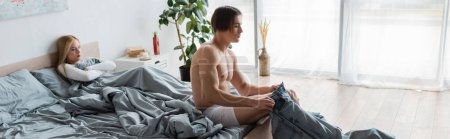 Téléchargez les photos : Homme torse nu en sous-vêtements portant un jean près de femme mécontente au lit après une table de nuit, bannière - en image libre de droit