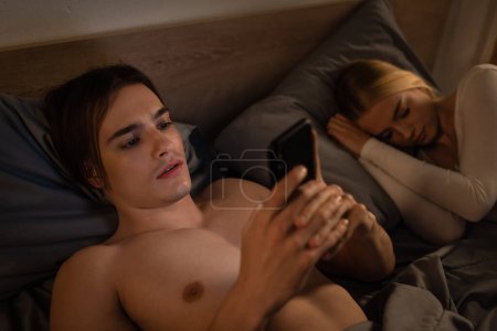 Foto de Hombre infiel mensajes de texto en el teléfono inteligente cerca de la novia durmiendo en la cama, concepto de engaño - Imagen libre de derechos