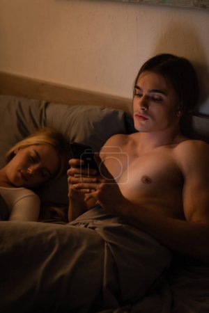hombre sin camisa usando el texto del teléfono móvil a la mujer rubia durmiendo en el dormitorio, concepto de engaño 