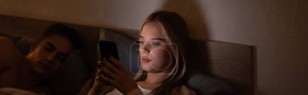 blonde woman messaging on smartphone next to sleeping boyfriend at night, banner 