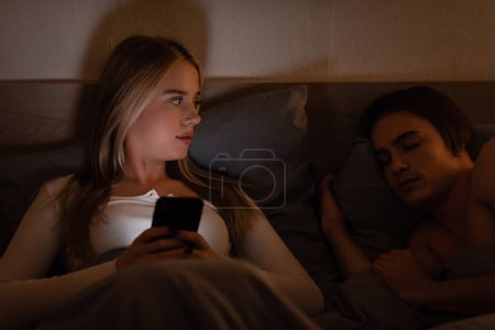 Téléchargez les photos : Femme blonde en utilisant smartphone à côté de dormir petit ami la nuit, concept de tricherie - en image libre de droit