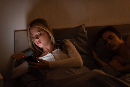 Téléchargez les photos : Femme infidèle en utilisant smartphone à côté de dormir petit ami la nuit, concept de tricherie - en image libre de droit