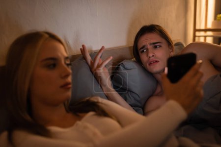 Photo for Displeased man gesturing while looking at upset girlfriend showing smartphone - Royalty Free Image