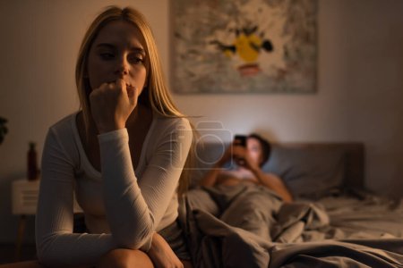 Besorgte Frau sitzt mit Smartphone im Schlafzimmer neben verschwommenem Freund im Bett 