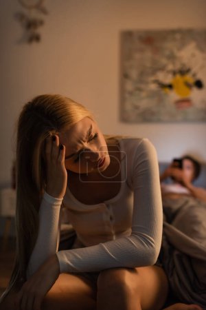 Verärgerte Frau sitzt neben verschwommenem Freund im Schlafzimmer auf Bett 