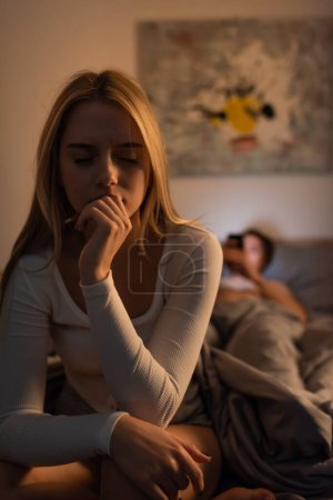 Besorgte Frau sitzt auf Bett neben verschwommenem Freund im Schlafzimmer und betrügt Konzept 