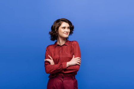 cheerful woman with short curly hair standing with folded arms isolated on blue 