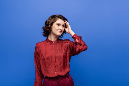 pensive language teacher in red blouse smiling while looking away isolated on blue 