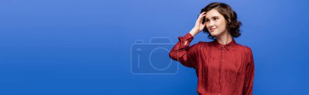 thoughtful language teacher in red blouse smiling while looking away isolated on blue, banner 