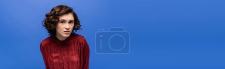 attentive language teacher in red blouse looking at camera isolated on blue, banner 