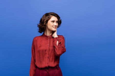 satisfied woman in red blouse with animal print smiling while looking away isolated on blue 