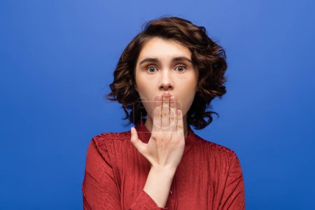 Photo for Shocked language teacher covering mouth with hand while looking at camera isolated on blue - Royalty Free Image