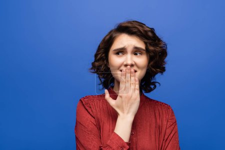 Photo for Nervous language teacher covering mouth with hand while looking away isolated on blue - Royalty Free Image