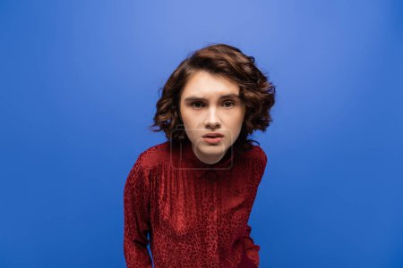 curious language teacher with curly short hair looking at camera isolated on blue 