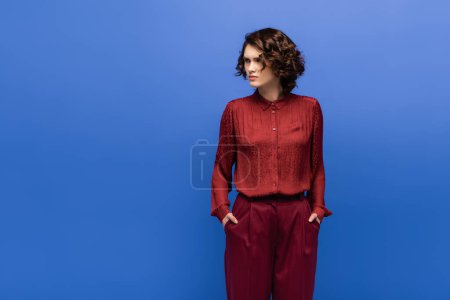 displeased language teacher in maroon color pants standing with hands in pockets isolated on blue 