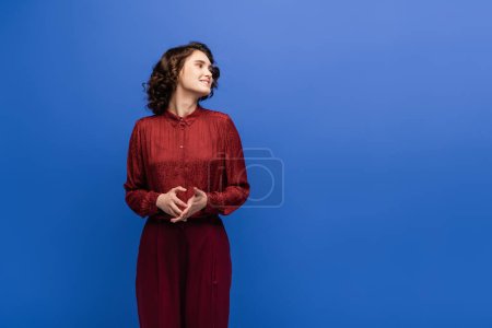 cheerful language teacher in maroon color outfit smiling while looking away isolated on blue 