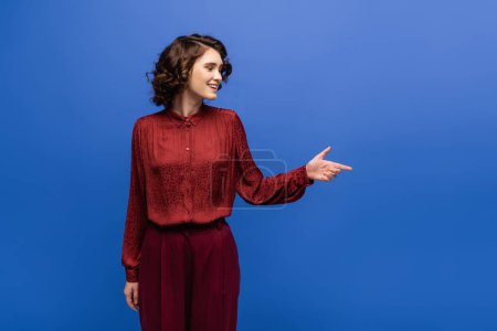 cheerful language teacher in maroon color outfit smiling while pointing with finger isolated on blue 