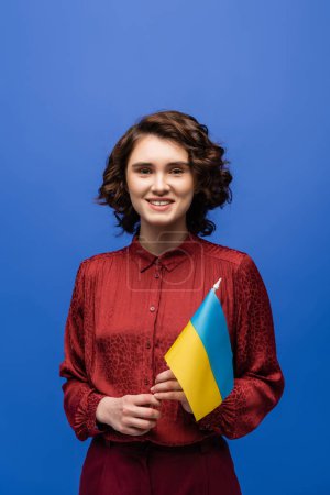 happy language teacher holding flag of Ukraine and smiling isolated on blue 