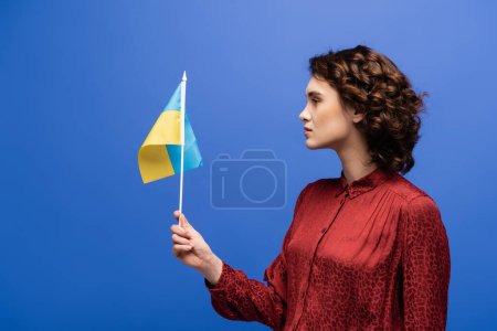 Photo pour Vue latérale du professeur de langue bouclée tenant le drapeau de l'Ukraine isolé sur bleu - image libre de droit