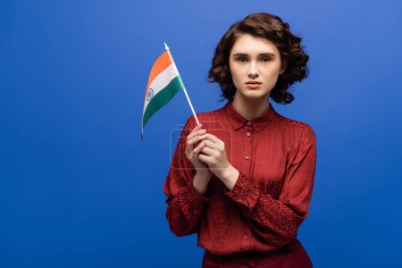 curly language teacher holding flag of India and looking at camera isolated on blue 