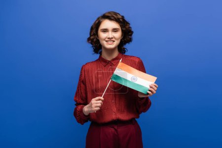 glückliche Frau lächelt, während sie die Flagge Indiens in die Kamera hält und isoliert auf blau blickt 