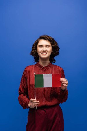 glückliche Sprachlehrerin lächelt, während sie Italien-Fahne in die Kamera hält und isoliert auf blau blickt 