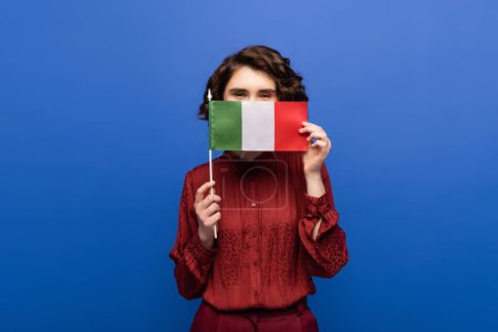 curly language teacher covering face while holding flag of Italy and looking at camera isolated on blue 
