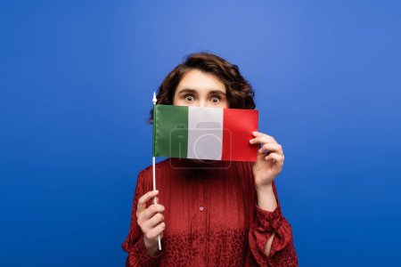 jeune femme bouclée couvrant le visage tout en tenant le drapeau de l'Italie et en regardant la caméra isolée sur bleu 