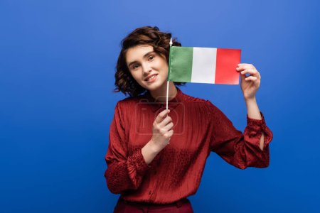 glückliche Frau lächelt, während sie die Flagge Italiens in die Kamera hält und isoliert auf blau blickt 