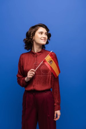 happy language teacher smiling while holding flag of Spain isolated on blue 