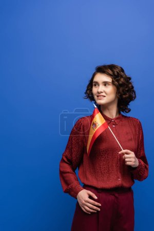 cheerful language teacher smiling while holding flag of Spain isolated on blue 
