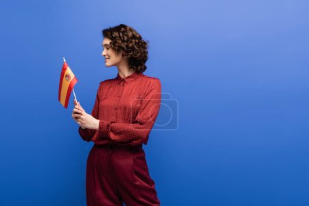 happy language teacher with curly hair holding flag of Spain isolated on blue 