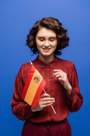 happy language teacher smiling while looking at flag of Spain isolated on blue 