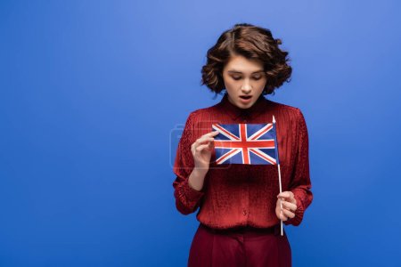shocked student with curly hair looking at flag of United Kingdom isolated on blue 