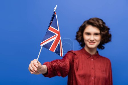 happy student with curly hair looking at flag of United Kingdom isolated on blue 