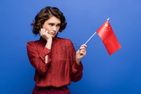 displeased student with curly hair looking at flag of China isolated on blue 