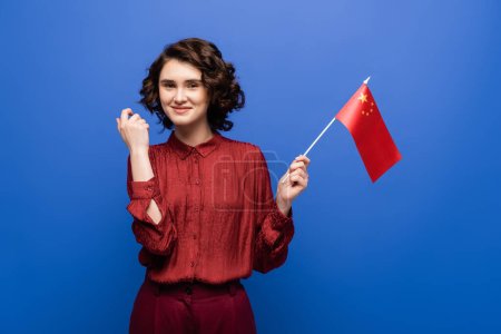 happy young student with curly hair looking at flag of China isolated on blue 
