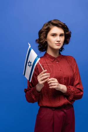 junge und ernsthafte Sprachlehrerin mit israelischer Fahne, die isoliert auf blauem Grund in die Kamera blickt