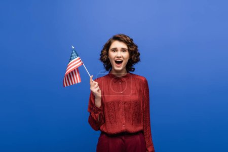 mujer emocionada con la boca abierta sosteniendo la bandera de EE.UU. y mirando a la cámara aislada en azul