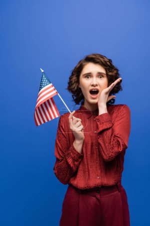 impactado estudiante con la boca abierta sosteniendo bandera de EE.UU. y mirando a la cámara aislada en azul