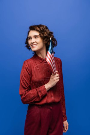 Photo for Brunette language teacher in burgundy blouse looking away while holding usa flag isolated on blue - Royalty Free Image