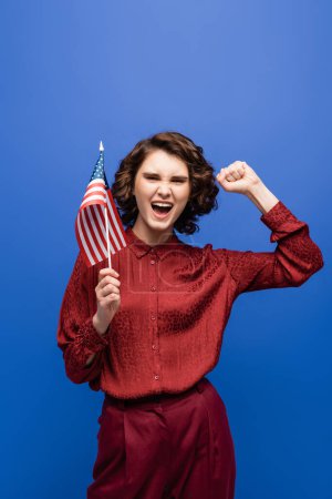 estudiante emocionado mostrando el gesto de éxito mientras sostiene la bandera de EE.UU. y mirando a la cámara aislada en azul