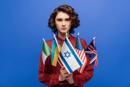 young and confident woman looking at camera while holding international flags isolated on blue