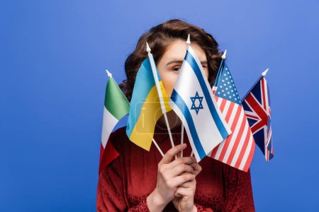 young woman looking at camera behind various international flags isolated on blue