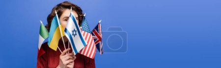 jeune femme obscurcissant le visage avec différents drapeaux et regardant la caméra isolée sur bleu, bannière