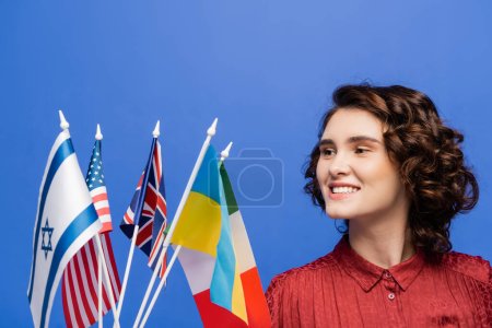 pleased young woman looking at flags of various countries isolated on blue